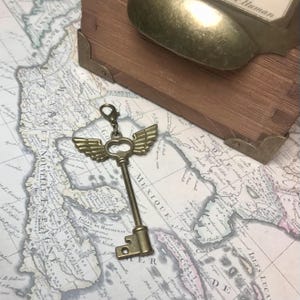 May include: A bronze-colored key pendant with attached wings and a heart-shaped detail. The key is displayed on a vintage map, with a wooden box and a spoon in the background. The key is a decorative item.