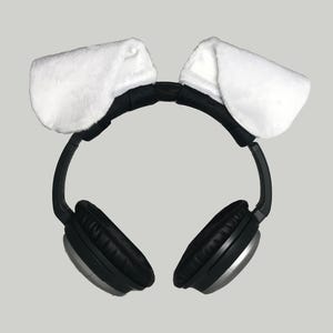May include: Black headphones with silver accents and plush white dog ears attached to the headband. The ear covers are black and the headband is black. The dog ears are made of a soft, white material.