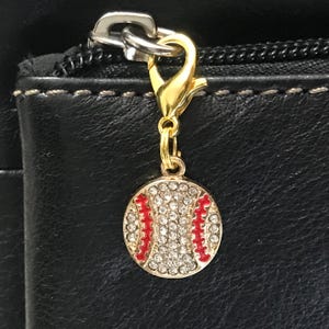 May include: A gold-colored baseball charm with a lobster clasp. The charm is round, covered in clear rhinestones, and features red stitching details. The charm is attached to a black leather surface.