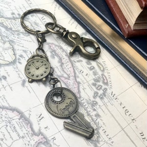 May include: Antique bronze-toned keychain featuring a small alarm clock charm and a key charm with the word "Empire" on it. The keychain includes a clasp and a key ring. The background includes a map and a leather-bound book.
