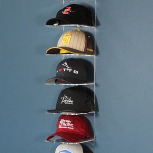 Könnte beinhalten: Ein klarer, an der Wand montierter Hutständer aus Acryl, der sieben Baseballkappen zeigt. Die Kappen sind schwarz, grau und rot, mit verschiedenen Logos und Designs. Der Ständer ist an einer blauen Wand befestigt.