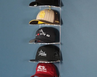 May include: A clear acrylic wall-mounted hat rack displaying seven baseball caps. The caps are black, gray, and red, with various logos and designs. The rack is mounted on a blue wall.