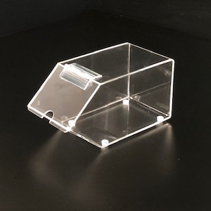 May include: A clear acrylic dispenser with a hinged lid. The dispenser has a small lip on the front for dispensing items.