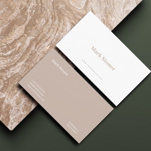 May include: Two business cards with the name "Mark Stoner" printed on them. One card is white and the other is a light brown color. The cards are on a green and brown textured surface.
