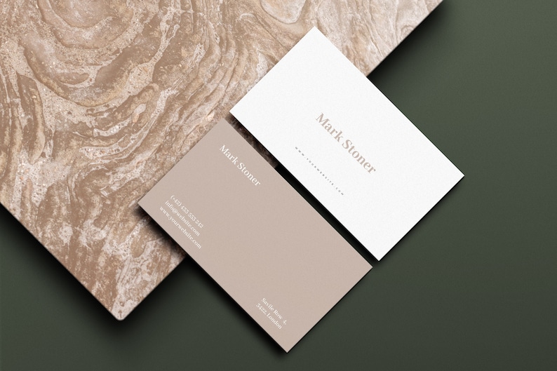 May include: Two business cards with the name "Mark Stoner" printed on them. One card is white and the other is a light brown color. The cards are on a green and brown textured surface.