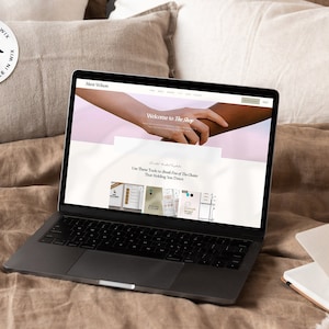 Puede incluir: Un port&aacute;til que muestra un sitio web con el texto "Welcome to The Shop" y un logotipo de "Wix". El port&aacute;til est&aacute; abierto sobre una cama con almohadas y un cuaderno.