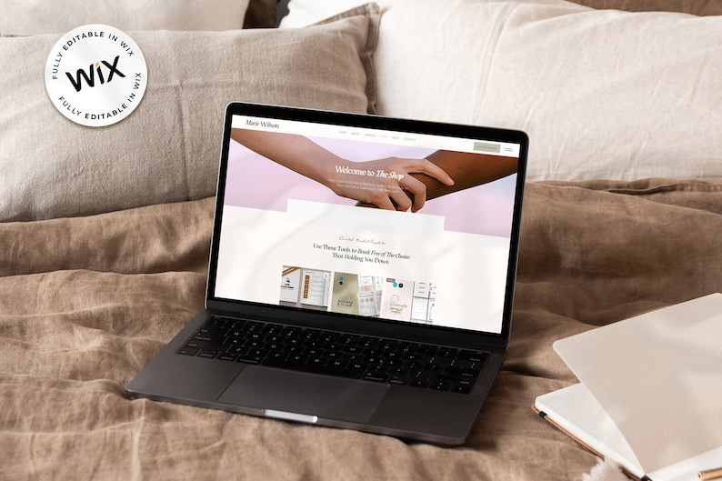 Puede incluir: Un port&aacute;til que muestra un sitio web con el texto "Welcome to The Shop" y un logotipo de "Wix". El port&aacute;til est&aacute; abierto sobre una cama con almohadas y un cuaderno.