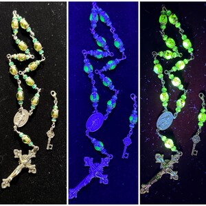 May include: A green and gold rosary with a silver crucifix and a silver medal of the Virgin Mary. The rosary beads glow green under a black light.