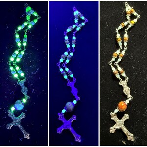 May include: A silver rosary with a cross pendant. The beads are made of a variety of materials, including wood, glass, and metal. The rosary is shown in three different lighting conditions, including normal light, UV light, and blacklight. The beads glow in the dark.