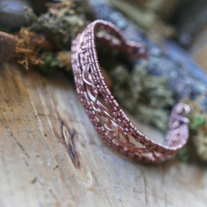 May include: A copper-colored wire-wrapped cuff bracelet. The openwork design features intricate weaving and a textured surface. The bracelet is displayed on a wooden surface with moss and a blurred background.