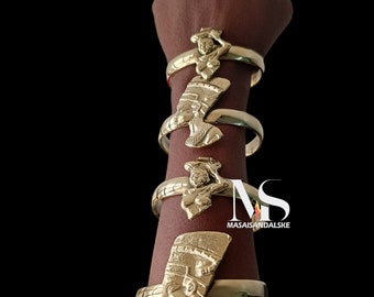 May include: Four gold bracelets with different Egyptian-inspired designs. Each bracelet features a raised image of a woman's head with a headdress. The bracelets are stacked on a person's arm. The text "MASAISANDALSKE" is visible on the bottom right corner of the image.