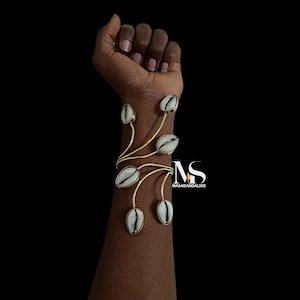May include: A gold metal bracelet with five white cowrie shells attached. The bracelet is on a person's wrist.