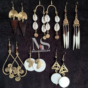 Puede incluir: Conjunto de seis pares de pendientes de tono dorado con varios diseños, incluyendo conchas, espirales y formas geométricas. Algunos pendientes presentan detalles en blanco y negro. Los pendientes se muestran sobre un fondo negro con el texto "MASAIHANDALSKE".