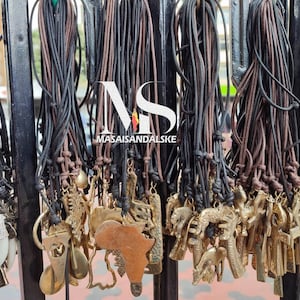 May include: A display of necklaces with various pendant designs, including an African continent shape, musical notes, and animal figures. The necklaces feature black and brown cords with metal charms. The text "MASAISANDALSKE" is visible in the image.
