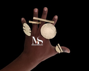 May include: A hand displays several gold-colored rings with unique designs. The rings include a textured circle, a leaf shape, a bar, and a half-circle. The background is solid black. The rings are likely fashion accessories.