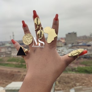 May include: A hand displaying several gold-colored rings. The rings feature various designs, including a triangle, a coin-like shape, a map of Africa, and shell-like forms. The nails are painted red.