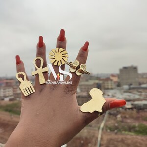 Pode incluir: Uma mão usando cinco anéis de ouro. Os anéis são em forma de sol, ankh, pente afro, borboleta e o contorno de um país. Os anéis estão nos dedos de uma pessoa com esmalte de unha vermelho.