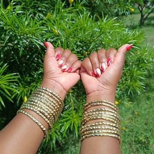 Pu&ograve; includere: Braccialetti color oro con una trama martellata sono impilati su entrambi i polsi. Le unghie sono dipinte con un disegno rosso e bianco. Lo sfondo &egrave; un verde lussureggiante con fiori gialli.