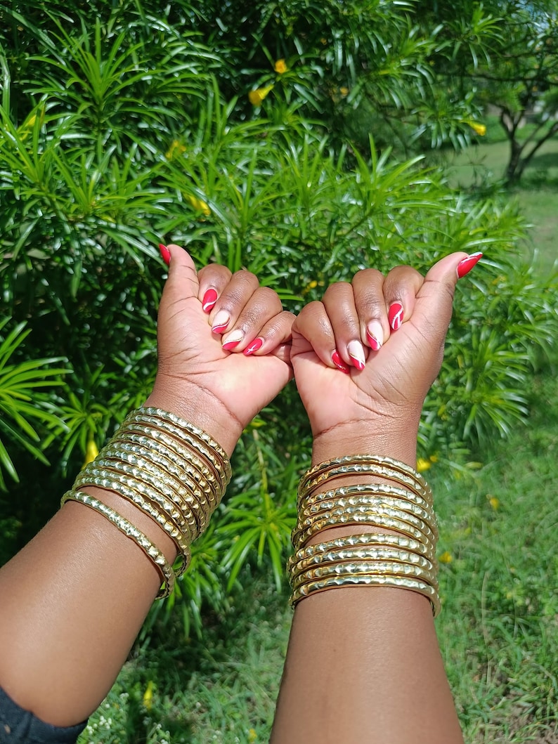 Pu&ograve; includere: Braccialetti color oro con una trama martellata sono impilati su entrambi i polsi. Le unghie sono dipinte con un disegno rosso e bianco. Lo sfondo &egrave; un verde lussureggiante con fiori gialli.