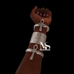 May include: Silver metal bracelets with Ankh symbols. The image shows three bracelets on an arm, one wide cuff and two thinner bands. The background is black. The brand name "MASAISANDALSKE" is visible.