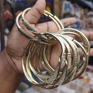 May include: A set of gold-toned metal bangles featuring a textured, woven-style pattern across the surface. The bracelets are circular with a polished finish and a repeating cross-hatched design, creating a consistent look across each individual piece.
