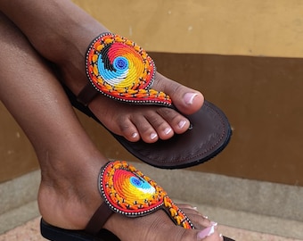 Könnte beinhalten: Braune Ledersandalen mit einem farbenfrohen Perlendesign. Die Sandalen weisen ein Spiralmuster in Rot-, Orange-, Gelb-, Blau- und Schwarztönen auf. Die Sandalen haben einen Zehensteg und werden an den Füßen getragen.