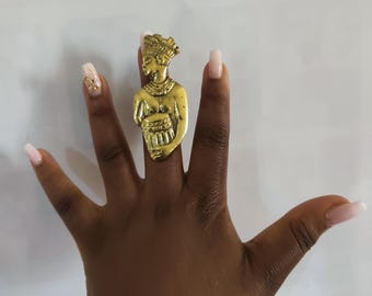 May include: A gold-colored ring featuring a detailed bust of a figure wearing a crown and holding an object. The ring is worn on a finger, showcasing the intricate design and craftsmanship. The ring's color contrasts with the skin tone.