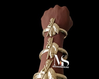 May include: Three gold bracelets with a warrior figure design. Each bracelet features a detailed warrior figure holding a spear and shield. The bracelets are stacked on a person's arm.