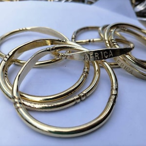 Peut inclure: Un ensemble de bracelets en métal couleur or avec le mot "AFRICA" gravé sur l'un des bracelets.