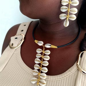 May include: A necklace and earrings set featuring white seashells and gold-coloured wire. The necklace has a black cord and the earrings dangle. The jewellery is displayed on a person wearing a beige top.