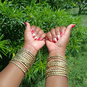 Pu&ograve; includere: Due braccia adornate da multipli braccialetti color oro con superficie strutturata. Lo sfondo &egrave; un verde lussureggiante con fogliame e fiori gialli. Le unghie sono dipinte di rosso e bianco.