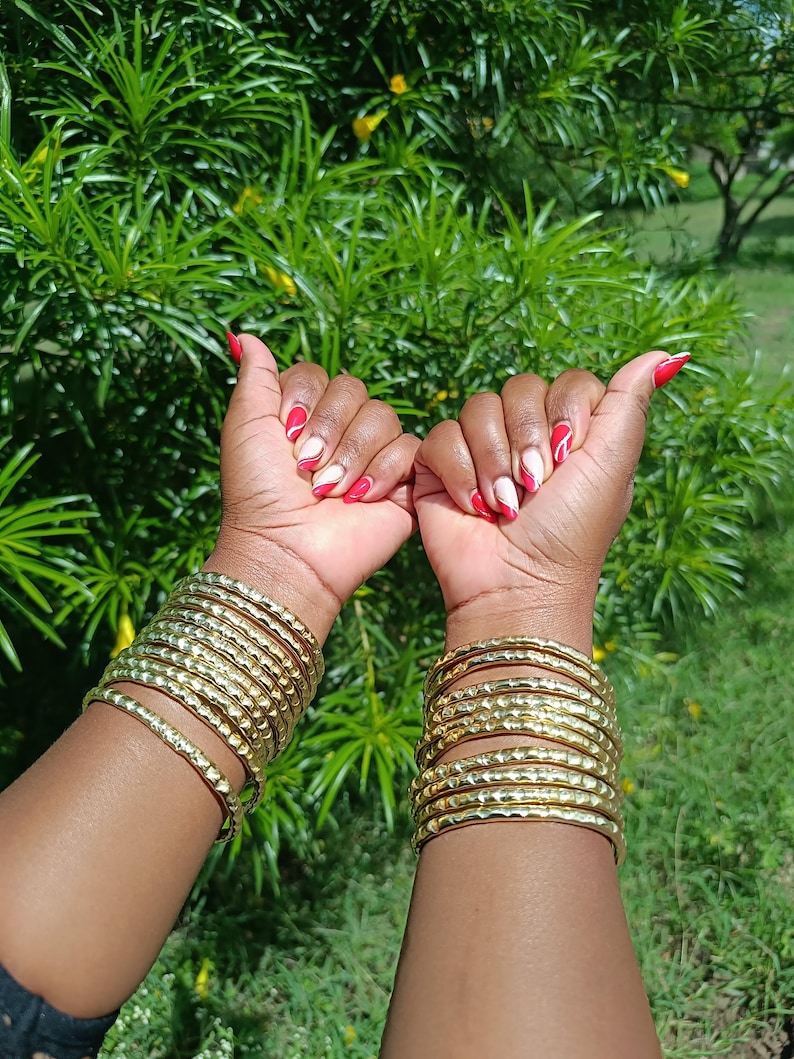Pu&ograve; includere: Due braccia adornate da multipli braccialetti color oro con superficie strutturata. Lo sfondo &egrave; un verde lussureggiante con fogliame e fiori gialli. Le unghie sono dipinte di rosso e bianco.