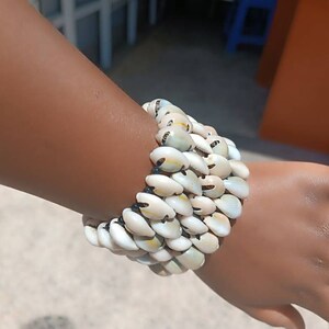 May include: A white and brown shell bracelet with multiple rows of shells. The bracelet is worn on a person's wrist.
