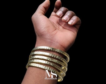 Puede incluir: Cinco pulseras de metal dorado apiladas en la muñeca de una persona. Las pulseras tienen un acabado texturizado y martillado.