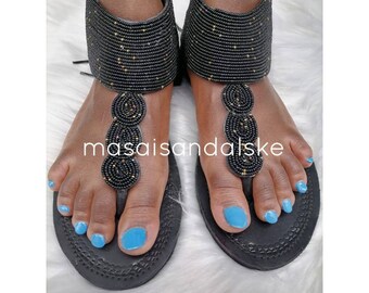 Könnte beinhalten: Schwarze Perlensandalen mit goldfarbenem Akzent. Die Sandalen haben ein T-Riemen-Design und einen Perlen-Knöchelriemen.