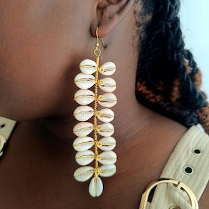 May include: Gold-coloured wire earrings with multiple white and cream-coloured seashells. The shells are arranged vertically, in a cascading design, with a single shell at the bottom. The earrings have a hook closure.