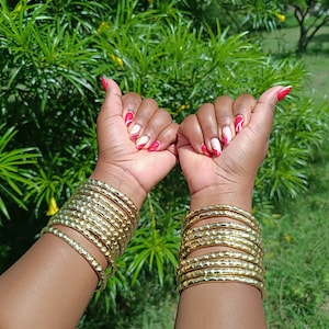 Pu&ograve; includere: Braccia adornate da multipli braccialetti color oro. Le unghie sono dipinte con un disegno rosso e bianco. Lo sfondo presenta un rigoglioso fogliame verde. I braccialetti sono in metallo.