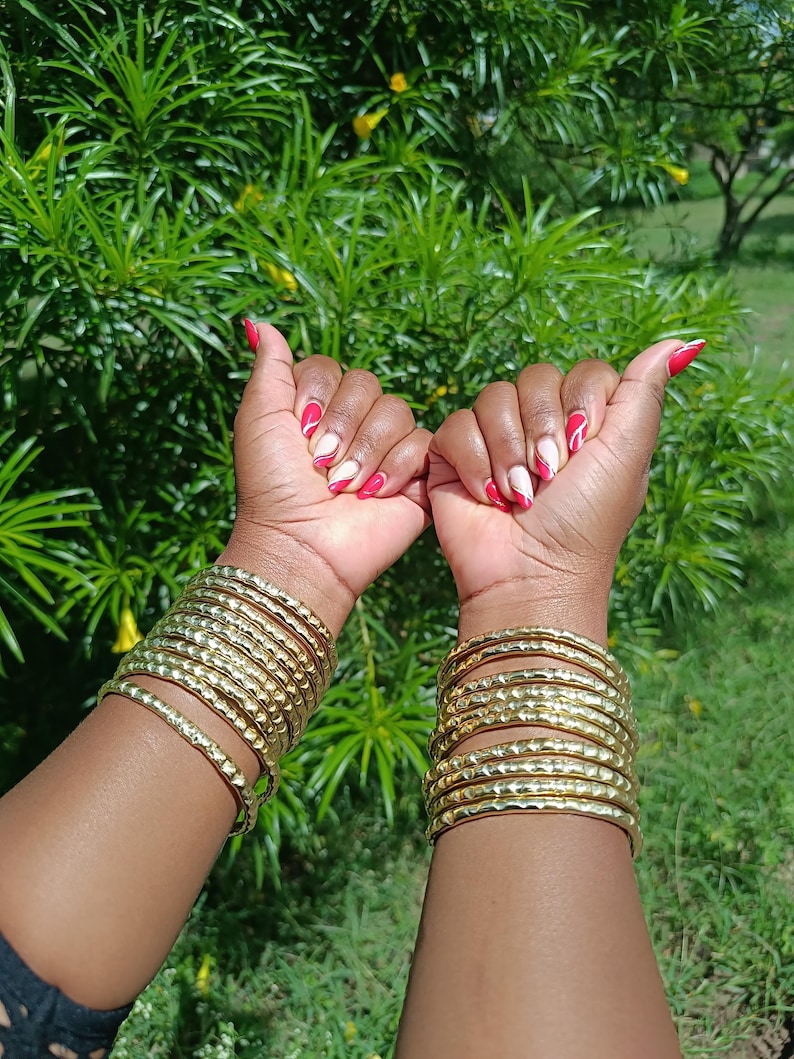 Pu&ograve; includere: Braccia adornate da multipli braccialetti color oro. Le unghie sono dipinte con un disegno rosso e bianco. Lo sfondo presenta un rigoglioso fogliame verde. I braccialetti sono in metallo.