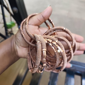Puede incluir: Una colección de pulseras de oro rosa. Las pulseras tienen un diseño de malla tejida y se sujetan con un cierre. Las pulseras son de un color metálico cálido y parecen estar hechas de metal.
