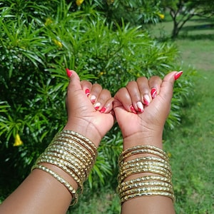 Pu&ograve; includere: Due braccia sono adornate da multipli braccialetti d'oro. I braccialetti hanno una superficie strutturata. Le unghie sono dipinte con un disegno rosso e bianco. Lo sfondo &egrave; un verde lussureggiante con fogliame.