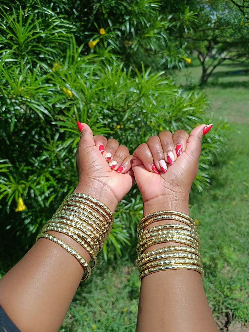 Pu&ograve; includere: Due braccia sono adornate da multipli braccialetti d'oro. I braccialetti hanno una superficie strutturata. Le unghie sono dipinte con un disegno rosso e bianco. Lo sfondo &egrave; un verde lussureggiante con fogliame.