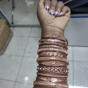 Peut inclure: Une collection de dix bracelets en métal de couleur cuivre empilés sur un bras. Les bracelets présentent diverses textures, notamment des motifs tressés, torsadés, nervurés et lisses, avec certaines pièces aux extrémités superposées.