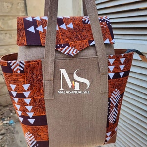 Op de afbeelding: Een bruine canvas tote bag met oranje en witte geometrische stoffen accenten. De tas heeft twee lange hengsels en een ritsvak aan de voorkant. De tas heeft de tekst "MASAISANDALSKE" op de voorkant gedrukt.