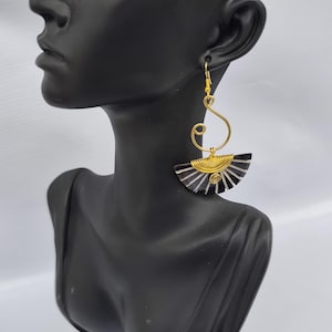 Può includere: Un paio di orecchini color oro con un design a forma di ventaglio. Gli orecchini presentano un motivo a righe bianche e nere con una spirale dorata al centro. Gli orecchini sono esposti su una testa di manichino nera.