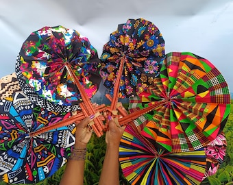 Könnte beinhalten: Eine Sammlung farbenfroher, gemusterter Handfächer. Die Fächer weisen lebendige Designs in verschiedenen Farben auf, darunter Rot, Grün, Blau und Gelb. Die Griffe bestehen aus einem rötlich-braunen Material. Die Fächer werden vor einem Hintergrund aus grünen Feldern und einem bewölkten Himmel gehalten.