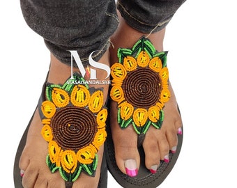 Könnte beinhalten: Ein Paar schwarze Ledersandalen mit Perlen-Sonnenblumen-Verzierungen. Die Sandalen haben einen Zehentrenner und sind mit gelben, orangefarbenen und grünen Perlen verziert.