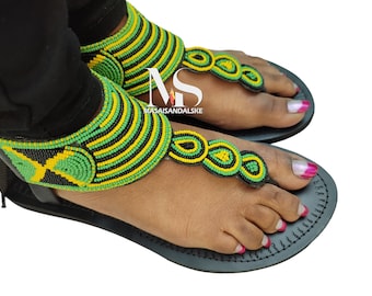 Könnte beinhalten: Schwarze Ledersandalen mit grünen, gelben und schwarzen Perlenriemen. Die Perlenriemen haben ein Design, das an die jamaikanische Flagge erinnert.