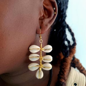Può includere: Orecchini dorati fatti a mano con un gruppo di conchiglie bianche. Le conchiglie sono disposte verticalmente in un design simmetrico, collegate da filo d'oro. Gli orecchini pendono da un gancio dorato.
