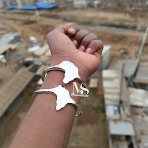 Puede incluir: Pulsera de plata con un diseño del continente africano. La pulsera se lleva en la muñeca de una persona. La marca "MASAISANDALSKE" es visible en la pulsera.