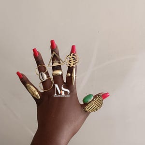 May include: A hand displaying a collection of gold-toned rings with various designs, including a triangle, a shell, and a leaf shape. The hand has red nail polish. The rings are of different sizes and styles, showcasing a variety of textures and shapes.